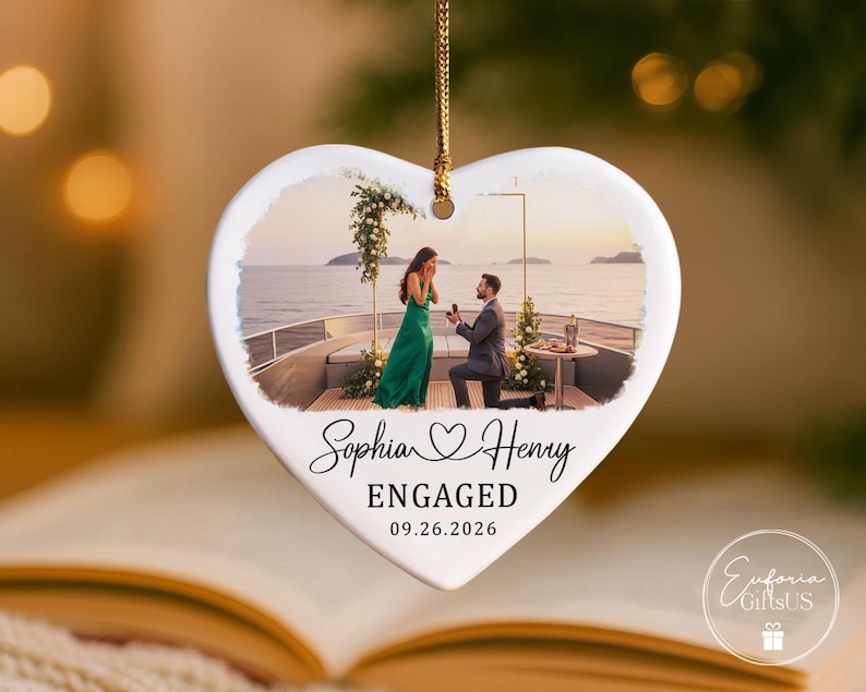 Puede incluir: Un adorno de cer&aacute;mica blanco en forma de coraz&oacute;n que cuelga de un cord&oacute;n dorado. El centro presenta una fotograf&iacute;a impresa de una propuesta de matrimonio en un barco. Debajo de la foto, el texto dice Sophia and Henry, ENGAGED, y la fecha 09.26.2026.