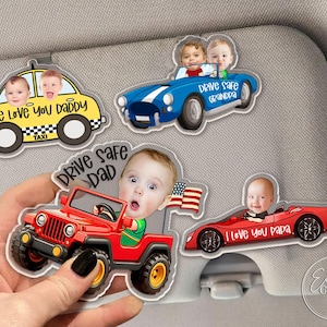 May include: A collection of car-shaped visor clips featuring baby faces. The clips include a yellow taxi with "We Love You Daddy", a blue convertible with "Drive Safe Grandpa", a red jeep with "Drive Safe Dad", and a red sports car with "I Love You Papa".