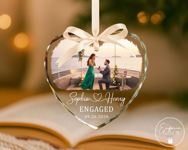 Puede incluir: Un adorno de cristal en forma de coraz&oacute;n con una fotograf&iacute;a impresa de una propuesta de matrimonio en la cubierta de un barco. El texto dice Sophia Henry, ENGAGED, y 09.26.2026. Una cinta de sat&eacute;n blanca forma un lazo para colgar el adorno.