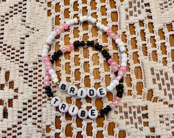 May include: Two beaded bracelets with the words "BRIDE" and "TRIBE" spelled out in white letters. The bracelets are made with white, pink, and black beads.