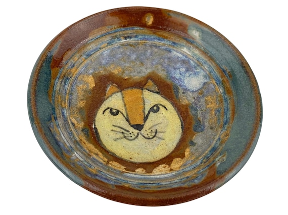 A hand-thrown stoneware plate with a sculpted lion face and a moody blue-green glaze — the kind of one-off ceramic you find at a gallery, not a craft fair.
