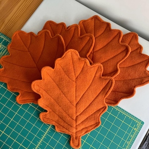 May include: A set of four orange fabric leaf-shaped placemats with stitched vein details. Each placemat features a scalloped edge and a soft, textured surface. The leaves are arranged in a cluster, showcasing their uniform colour and shape.
