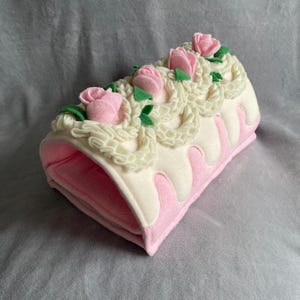 Peut inclure: Un rouleau en feutrine cylindrique décoré pour ressembler à un gâteau avec des roses en feutrine rose, des volutes de glaçage blanc et des feuilles vertes. La base est rose clair avec un glaçage blanc qui coule sur les côtés.
