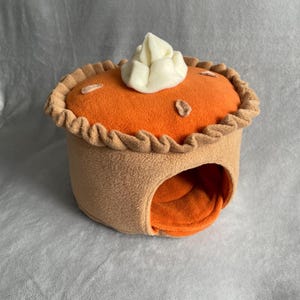 May include: A plush pet bed shaped like a pumpkin pie with a tan base, an orange top, a ruffled crust edge, and a white dollop of cream on top. The front features a circular opening leading to an orange interior, crafted from soft fabric.