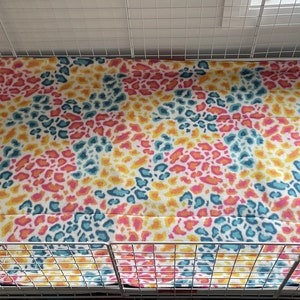 May include: A rectangular pet cage liner featuring a vibrant animal print pattern with overlapping spots in shades of bright yellow, hot pink, and teal blue. The fabric is laid flat inside a white wire grid enclosure with a solid white base tray.