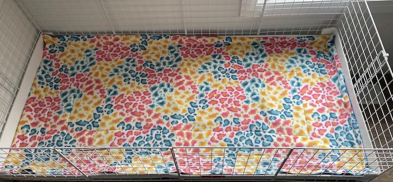 May include: A rectangular pet cage liner featuring a vibrant animal print pattern with overlapping spots in shades of bright yellow, hot pink, and teal blue. The fabric is laid flat inside a white wire grid enclosure with a solid white base tray.