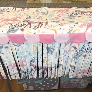 May include: A fleece fabric hideout featuring a white background with a pattern of pink and purple unicorns, yellow crescent moons, and blue foliage. The top edge has a row of pink scalloped fabric with long, hanging white fringe strips.