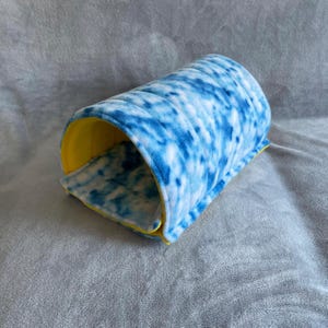 Peut inclure: Un lit pour animal en forme de tunnel avec un extérieur en peluche douce arborant un motif tie-dye bleu et blanc. L'intérieur du tunnel est doublé d'un tissu jaune vif uni, créant un contraste entre la surface extérieure et la doublure.