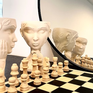 Könnte beinhalten: Ein weißes Schachbrett mit weißen Schachfiguren darauf. Im Hintergrund befinden sich drei weiße Keramik-Blumentöpfe mit Gesichtern, die in einem Spiegel reflektiert werden.