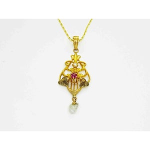May include: A gold-colored pendant necklace with an ornate design. The pendant features a central red gemstone, a small pearl, and decorative elements. The necklace is suspended from a delicate gold chain.