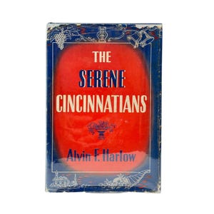 May include: A hardcover book titled The Serene Cincinnatians by Alvin F. Harlow. The cover features a vibrant red central panel framed by a dark blue border with white line art illustrations of grapes, bells, and a cornucopia at the bottom.