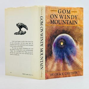 May include: A hardcover book titled "Gom on Windy Mountain" by Grace Chetwin. The cover features a stylized eye design with radiating lines in shades of purple, orange, and brown. The spine displays the title and author's name.