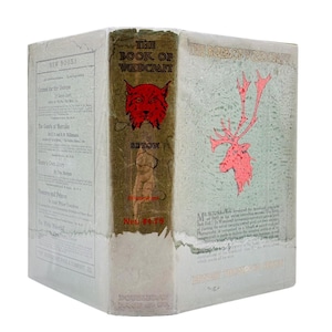 May include: A vintage book titled "The Book of Woodcraft" with a red bobcat illustration on the spine. The cover features a red deer illustration and text. The book is wrapped in a protective cover.