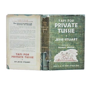 May include: An open book titled "Taps for Private Tussie" by Jesse Stuart. The cover is green with white text and an illustration. The book is a winner of the 1943 Thomas Jefferson Award and was a Book-of-the-Month Club selection.