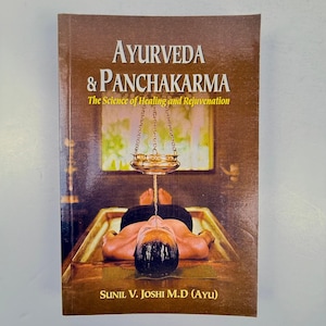 May include: A book cover with a brown background and the title "Ayurveda & Panchakarma: The Science of Healing and Rejuvenation" in gold lettering. The cover features a photo of a person receiving a traditional Ayurvedic treatment, with a metal pot hanging above their head and a stream of liquid pouring onto their forehead. The author's name, "Sunil V. Joshi M.D (AYU)", is printed in gold at the bottom of the cover.