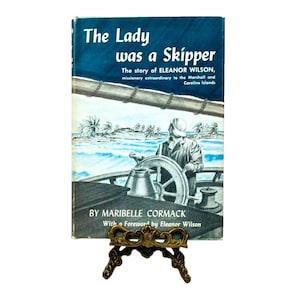 May include: Vintage book titled "The Lady was a Skipper" on a decorative brass stand. The cover features a blue sky, ocean scene, and a person at the helm of a ship. The book tells the story of Eleanor Wilson, a missionary.