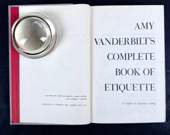 May include: An open book titled "Amy Vanderbilt's Complete Book of Etiquette" with a silver magnifying glass on the left page. The book has a red spine and the text "A Guide to Gracious Living".
