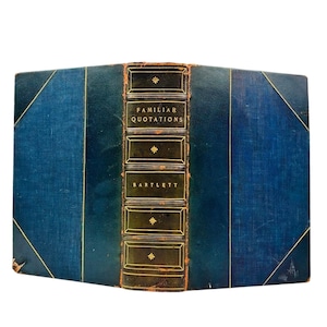 May include: Antique book titled "Familiar Quotations" by Bartlett. The book has a dark blue cover with gold lettering and accents. The spine features the title and author's name in gold rectangular frames. The book's edges are also blue.