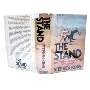 May include: A vintage copy of Stephen King's novel "The Stand." The book cover features a desert scene with figures in action, and the title is prominently displayed. The spine shows the title and author's name.