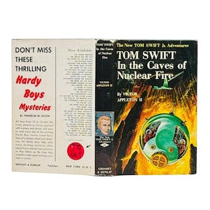 May include: A vintage book cover featuring "Hardy Boys Mysteries" and "Tom Swift in the Caves of Nuclear Fire." The cover has a retro design with bold text and illustrations, including a portrait of a young man and a green submarine-like vehicle.