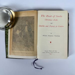 May include: A vintage book titled "The Book of Snobs" and "Christmas Books and Sketches and Travels in London" by William Makepeace Thackeray. The book is published by Thomas Nelson and Sons, London, Edinburgh, Dublin, and New York.