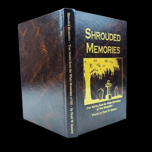 May include: A hardcover book titled "Shrouded Memories" with gold lettering and a yellow illustration of a tree and tombstones. The book's cover is dark brown with a textured appearance. The spine also features text.