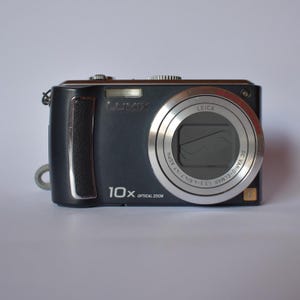 May include: A black Lumix digital camera with a silver lens and the text "LEICA" and "10X OPTICAL ZOOM". The camera has a built-in flash and a wrist strap attachment. The lens is surrounded by a silver ring.