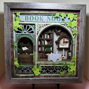 May include: A framed, multi-layered paper art piece depicting a cozy book nook. The artwork features a clock, bookshelves, and the text "BOOK NOOK" and "A BOOK IS A DREAM". Green ivy accents the design, with a rustic, wooden frame.