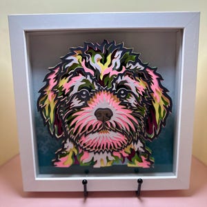 May include: A framed paper art piece of a dog's face. The dog is a white and pink color with black and green accents. The paper art is layered and has a 3D effect.