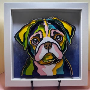 May include: A colorful paper cut-out of a pug dog's face in a white frame. The dog's fur is layered in shades of yellow, green, blue, and pink.
