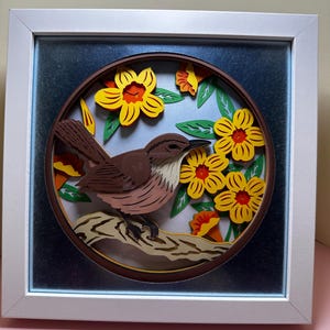 May include: A brown bird with a long tail perched on a branch surrounded by yellow flowers with orange centers. The artwork is framed in a white frame with a blue mat.