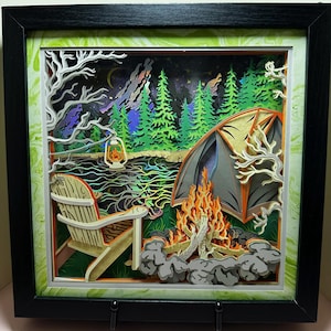 May include: A framed 3D paper art piece depicting a camping scene. The artwork features a campfire, tent, Adirondack chair, and a lantern near a lake, all under a starry night sky. The color palette includes greens, oranges, and blues, creating a layered effect.