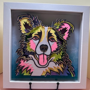 May include: A framed paper art piece of a dog's face. The dog is a border collie with a white face, black and green fur, and pink accents. The dog is smiling with its tongue sticking out.