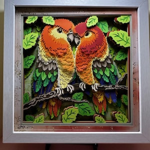 May include: A framed 3D paper art piece featuring two colorful parrots perched on a branch. The parrots have orange, red, green, and blue plumage, surrounded by green paper leaves. The artwork is in a silver frame.