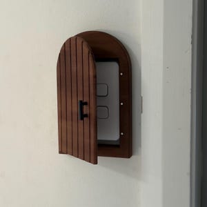May include: A wooden light switch cover in the shape of a small door, mounted on a white wall. The door is a dark brown with vertical grooves and a black handle. The light switch is visible behind the open door.