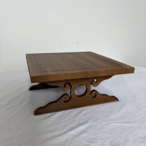 Op de afbeelding: Een kleine, vierkante houten tafel met een donkerbruine afwerking. De tafel heeft een decoratieve basis met gebogen uitsparingen en een verzonken bovenkant. De tafel is ongeveer 25 cm breed en 25 cm lang.