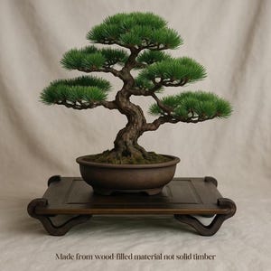 May include: A miniature bonsai tree with green leaves and a brown trunk, in a brown ceramic pot. The tree is on a dark wooden stand with curved legs. The text at the bottom reads: Made from wood-filled material not solid timber.