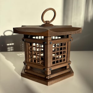 May include: A brown wooden lantern with a square base and a pagoda-style roof. The lantern has a grid design on its sides and a circular handle on top. The lantern is made of wood and has a dark brown finish.