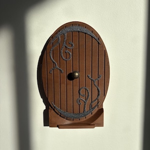 May include: A wooden, oval-shaped door with vertical planks and decorative black metal accents. The door features a brass doorknob and is mounted on a small wooden base. The design evokes a whimsical, fantasy theme.