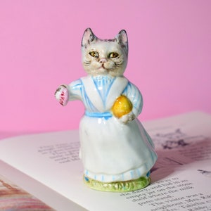 Puede incluir: Una figurita de porcelana de un gato con un vestido azul y blanco, sosteniendo un corazón rosa y un limón amarillo. La figurita está de pie sobre un libro abierto con texto.