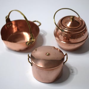 May include: Three small copper pots with brass handles and accents. One pot has a handle, another has a lid and handle, and the third has a lid and two small handles. The pots are displayed on a white surface.