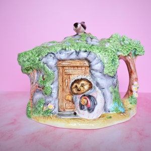 May include: A ceramic teapot shaped like a hedgehog's house with a small bird on the roof. The teapot is painted with green leaves, brown branches, and a grey stone exterior. A hedgehog is peeking out of the door.