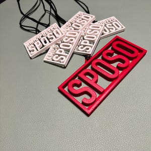 以下が含まれることがあります： 太字の「SPOSO」という文字が入った、白と赤の長方形のペンダント。白いペンダントには「AMICO DELL SPOSO」の文字も入っています。黒いコードが付いています。ペンダントは灰色の表面に置かれています。