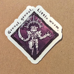 Puede incluir: Una pegatina blanca con una ilustración púrpura y negra de una figura robótica con una guadaña. El texto "Grovel, grovel, little worm." está escrito alrededor del borde de la pegatina.