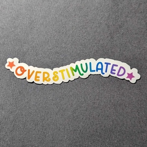 May include: A rainbow-colored sticker with the text "OVERStimulated" in a curved font with stars on either side.