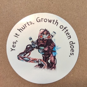 Puede incluir: Una pegatina redonda blanca con una ilustración en blanco y negro de un robot con ojos rojos. La pegatina tiene el texto "Sí, duele. El crecimiento a menudo lo hace." alrededor del borde.