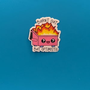 Puede incluir: Una pegatina rosa con un contenedor de basura de dibujos animados en llamas, con el texto "BURNT OUT" encima y "BUT OPTIMISTIC" debajo. El contenedor de basura tiene una cara sonriente y está sobre un fondo azul.