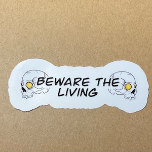 May include: White sticker with two black and white skull illustrations with yellow eyes. The text "BEWARE THE LIVING" is printed in black between the skulls.