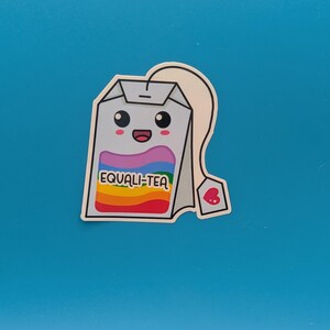 Può includere: Una bustina di tè di cartone animato con un design arcobaleno e il testo "EQUALI-TEA". La bustina di tè ha un viso sorridente e un'etichetta a forma di cuore.
