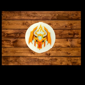 May include: A golden dragon head with flames and wings, painted on a white circle, against a brown wooden background.